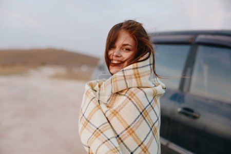 Téléchargez les photos : Femme profitant d'une expérience de voyage paisible dans le désert enveloppé dans une couverture à côté de la voiture - en image libre de droit