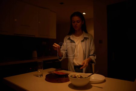 Femme préparant une collation en soirée dans une cuisine faiblement éclairée avec un bol de nourriture et une assiette devant elle