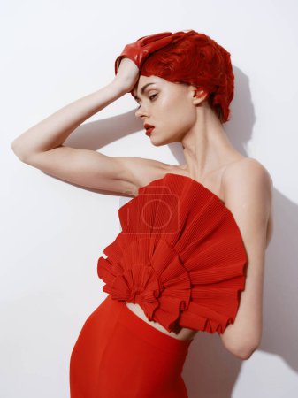 Schöne Frau mit rotem Kleid und roten Haaren posiert vor weißer Wand in eleganter Mode-Pose