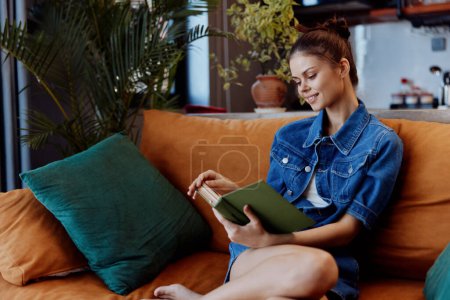 Young woman sitting on a couch leisurely reading a book in denim shirt and jeans in cozy home atmosphere