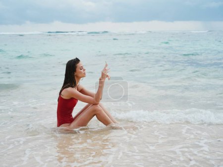 Téléchargez les photos : Femme avec un beau touriste bronzé dans un maillot de bain rouge assis sur le sable sur la plage dans l'océan dans les vagues coûteuses. Photo de haute qualité - en image libre de droit