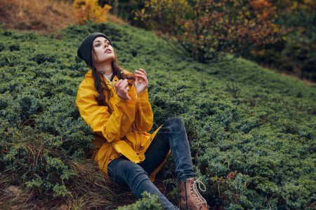 Jeune femme dans un imperméable jaune assis sur une colline herbeuse avec les mains dans les poches, profiter de la nature et des loisirs