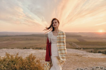 Frau in weißem Kleid und kariertem Schal steht bei Sonnenuntergang auf einem Hügel mit Reise- und Schönheitsthema