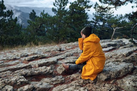 Einsamer Reisender in gelbem Regenmantel und Hut auf dem Gipfel des Berges sitzend und den atemberaubenden Horizont bewundernd