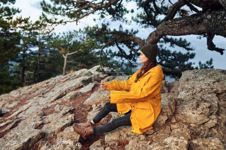 A woman in a yellow coat enjoys the scenic view from a rocky hilltop with a distant tree