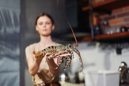 Woman holding a large lobster in a kitchen setting with a sink in the background, preparing seafood dish