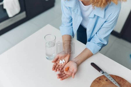 Woman taking medication in a kitchen setting with pills and a glass of water on a white table