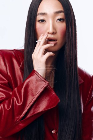 Mujer rebelde con el pelo largo negro y chaqueta de cuero rojo fumar cigarrillo, aislado sobre fondo blanco