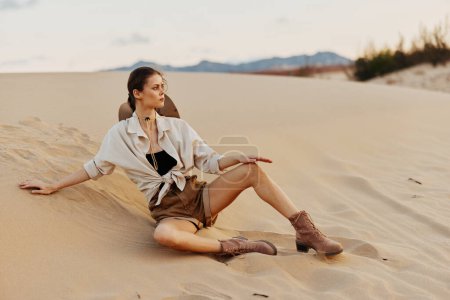 Femme reposant sur une dune sablonneuse avec chemise blanche et bottes brunes, profitant d'un moment paisible dans un paysage désertique