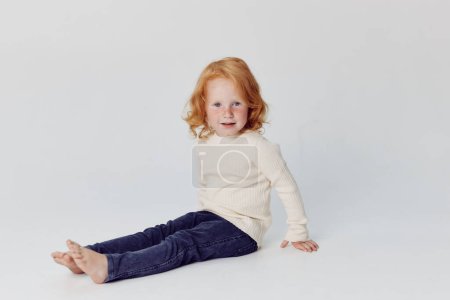 Adorable little girl with red hair sitting in front of white background, childhood beauty and innocence