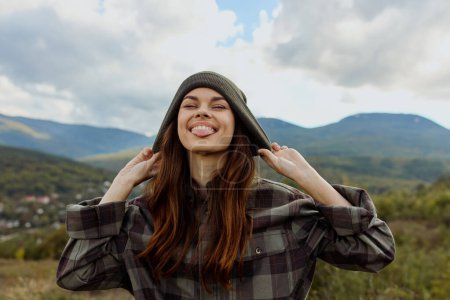 A stylish woman in a plaid shirt and hat posing against the majestic backdrop of a mountain range