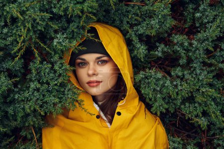 Eine junge Frau in einem gelben Regenmantel versteckt sich hinter immergrünen Bäumen in einer ruhigen Waldkulisse