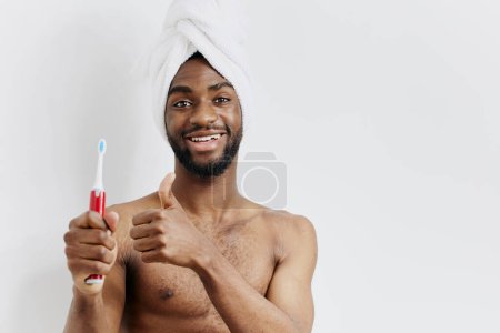 Hombre alegre en el baño con toalla envuelta en la cabeza, sosteniendo el cepillo de dientes y dando un pulgar confiado hacia arriba.
