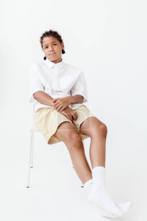 Stylish young boy in white shirt and shorts sits confidently with legs crossed on chair, exuding a fashionable and poised demeanor.