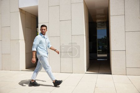 Ein selbstbewusster junger Mann in hellblauer Freizeitkleidung schreitet zielstrebig an einem modernen Gebäude vorbei, das urbanen Lebensstil und Ehrgeiz verkörpert.