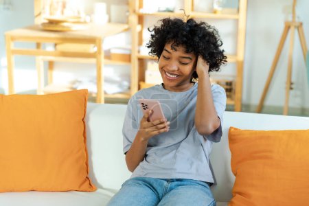 Aufgeregt glücklich junge schwarzafrikanische Amerikanerin hält Handy lachend Gefühl Freude bekommen mobile Nachricht. Überglückliches Mädchen, das laut lacht, wenn es auf der Couch sitzt und lustige Videos anschaut, in denen Nachrichten gelesen werden
