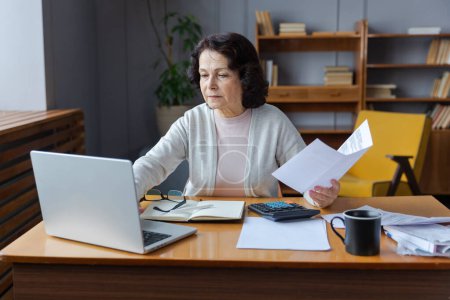 Middle aged senior woman sit with laptop and paper document. Pensive older mature lady reading paper bill pay online at home managing bank finances calculating taxes planning loan debt pension payment
