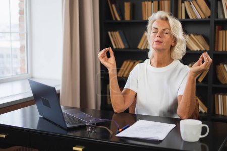 Yoga mindfulness meditation No stress keep calm. Middle aged woman practicing yoga at office. Woman taking break from work meditating relaxing. Mature lady doing breathing practice online yoga at work