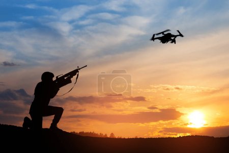 Téléchargez les photos : Soldat essayant d'abattre drone de reconnaissance dans le contexte d'un coucher de soleil. Un soldat tire sur un quadcopter. Méthodes modernes de guerre. Concept technologique. - en image libre de droit