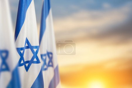 Foto de Israel flags with a star of David over cloudy sky background on sunset. Patriotic concept about Israel with national state symbols. Banner with place for text. - Imagen libre de derechos