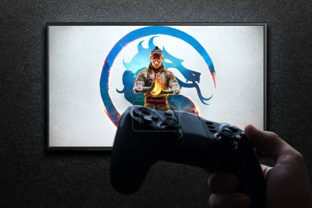 Foto de Mortal Kombat 1 juego en la pantalla de TV con mando en la mano en la pared de textura negro con luz. Astana, Kazajstán - 2 de julio de 2023. - Imagen libre de derechos