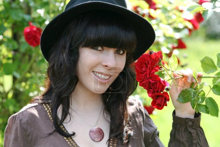 A smiling dark-haired young woman with a hat next to red roses