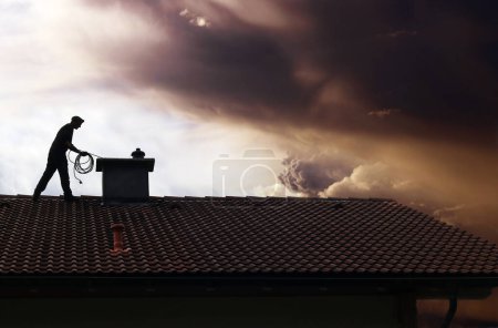 A chimney sweep cleans the chimney on a house roof while thunderclouds gather
