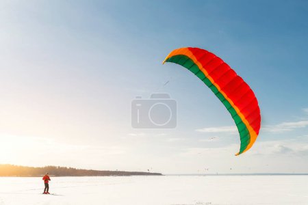 Panoramic view of many people friends enjoy riding kite surf board in warm suit on bright sunny winter day at frozen lake field snowy surface. Wintersport adrenaline fun adventure hobby acitivity.