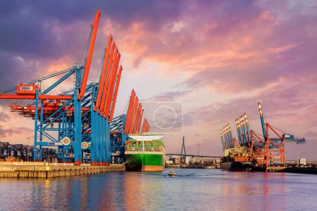 Scenic front giant cargo container ship loading Hamburg city port harbour seaport cranes warm dramatic sky evening sunset light. Global commercial trade freight charter shipping logistics background.