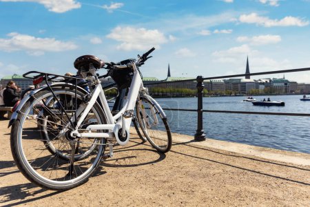 Photo for Scenic view two modern bicycles parked at embankment of Alster lake in Hamburg street old city center background summer day panorama view. Healthy travel lifestyle concept. Urban rental transport. - Royalty Free Image