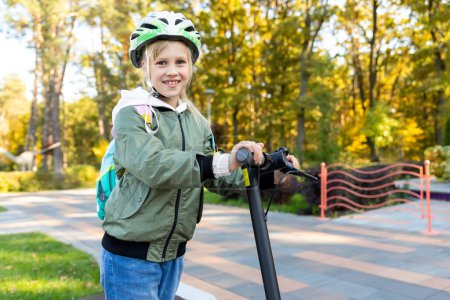 Photo for Profile view portrait of cute blond little caucasian school girl wear helmet enjoy having fun riding electric scooter city street park outdoors on sunny day. Healthy sport children activities outside. - Royalty Free Image