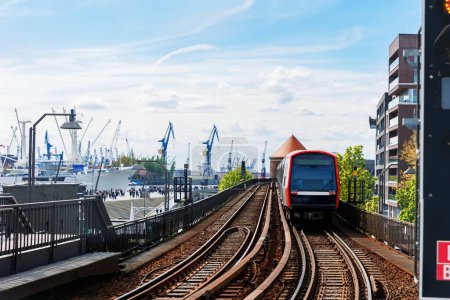 Hamburg Red U-bahn Subway train departure Baumwall station on Elevated Track with Hamburger Port Cranes and ships cargo harbor background blue sky. Hanseatic city commute transport scenic view.