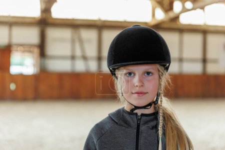 Portrait of cute confident young girl kid with braided hair wear riding helmet ready horseback riding lesson in sunlit arena hall stable farm ranch equestrian club school. Little beginner rider child.