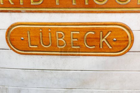 Foto detallada de una placa náutica en un barco, con la palabra LUBECK grabada en letras doradas sobre un fondo de madera pulida.