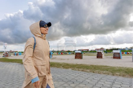 Smiling happy woman running by Nord sea coast wear beige coat  sunglasses enjoys windy day cloudy rainy stormy sky background. Female person have fun hiking adventure travel at Baltic sea seaside.