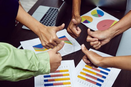 Photo for A collaborative team giving thumbs up over business performance charts on a table, indicating project approval and success - Royalty Free Image