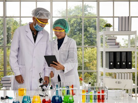 Two scientific in a bright lab examines results on a tablet, with one operating a microscope, surrounded by a variety of colorful chemical solutions