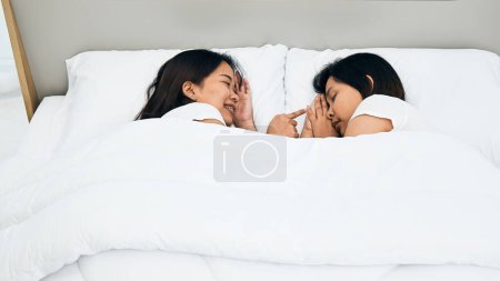 Asian young woman whispers playfully to her colleague as they enjoy a private moment together, nestled in a comfortable white bed