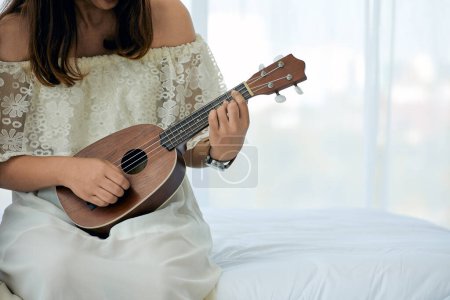 Gros plan des mains d'une femme jouant habilement de l'ukulélé, assise sur un lit blanc dans une chambre lumineuse, créant une mélodie apaisante