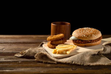 Photo for Hearty Burger meal on wood board with fried onion rings, fries, rustic appeal, dark background - Royalty Free Image