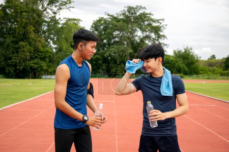 Two young Asian men cooling down, one wiping sweat with a towel, the other drinking water, after a workout on a running track