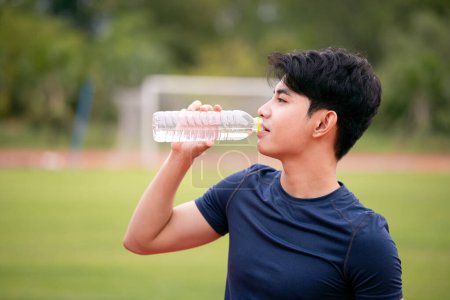 A young male athlete in sportswear quenches his thirst with a bottle of water on a sunny day at the athletic track