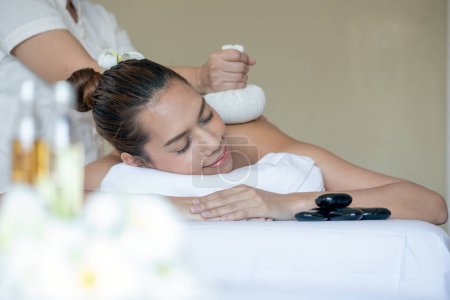 Peaceful atmosphere and young Asian woman enjoys soothing herbal compress massage at contemporary spa, surrounded by essential oils