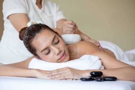 Asian young serene woman enjoys traditional herbal compress massage at spa, emphasizing relaxation and holistic wellness