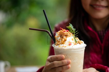 Primer plano de la mujer sosteniendo café helado gourmet cubierto con crema batida y granola, al aire libre en un entorno verde exuberante