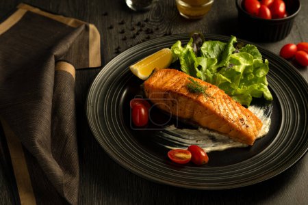 Grilled salmon fillet on dark circular plate, accompanied by fresh green salad, cherry tomatoes, and slice of lemon, on dark wooden table