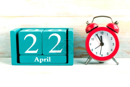 22 de abril. Calendario cubo azul con mes y fecha y reloj despertador rojo sobre fondo de madera