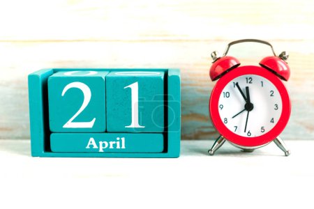 April 21. Blue cube calendar with month and date and red alarm clock on wooden backgroun