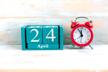 24 de abril. Calendario cubo azul con mes y fecha y despertador rojo sobre fondo de madera