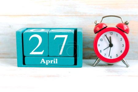April 27. Blue cube calendar with month and date and red alarm clock on wooden background